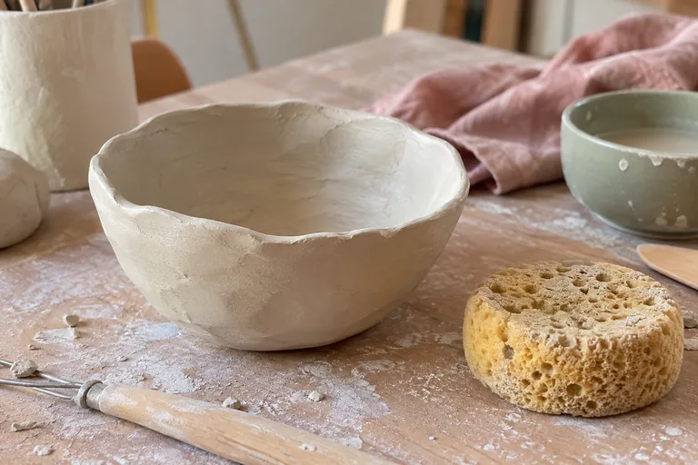 Bol d’argile aux bords irréguliers près d’une éponge sur une table d’atelier.