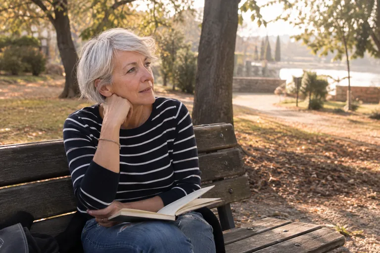 Une femme aux cheveux argentés, pensive sur un banc de parc, un carnet ouvert sur les genoux