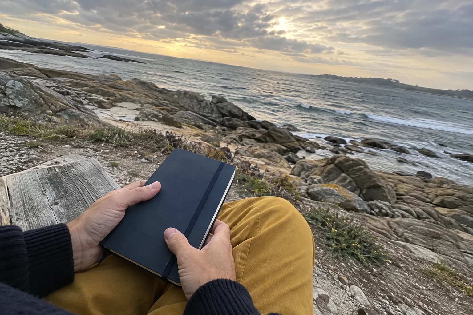 Des mains tiennent un carnet fermé sur un banc face à la mer