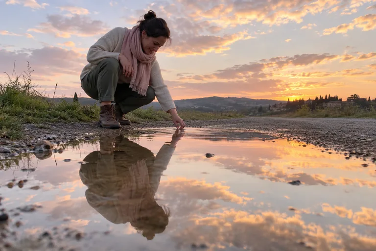 Une femme accroupie touche une flaque reflétant le ciel au soleil couchant.