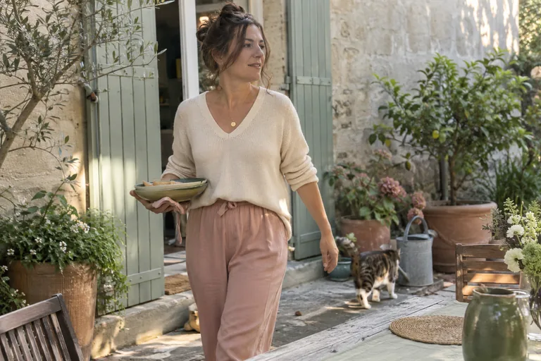Femme traversant une cour avec des assiettes, tandis qu’un chat passe derrière elle.
