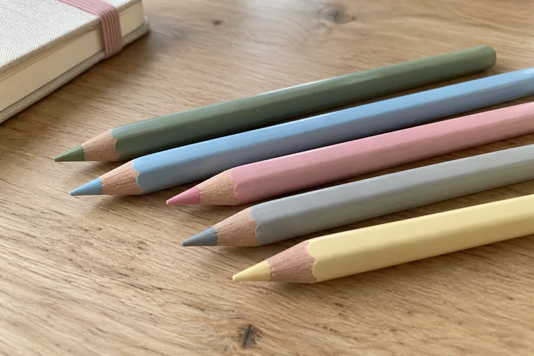 Cinq crayons vert, bleu, rose, gris et jaune pâle posés près d’un carnet fermé.