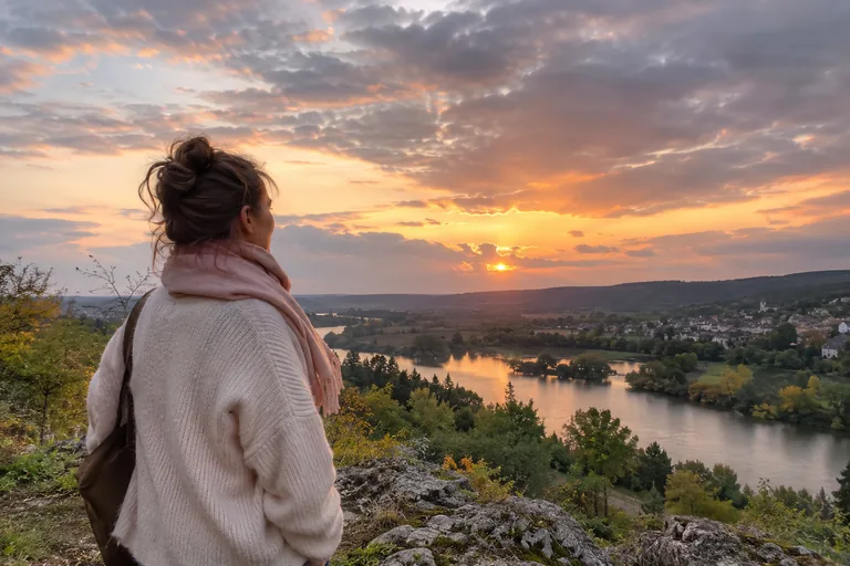 Femme regardant un coucher de soleil nuageux au-dessus d’une rivière et d’un village.
