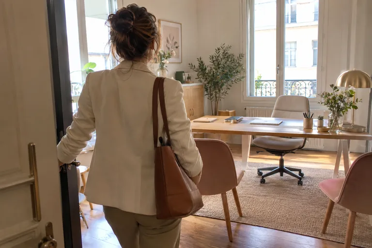 Femme vue de dos entrant dans un bureau lumineux avec un sac brun à l’épaule.
