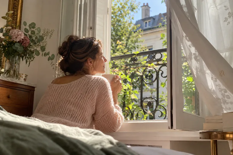 Femme tenant une tasse près d’une fenêtre ouverte sur des branches et des immeubles.