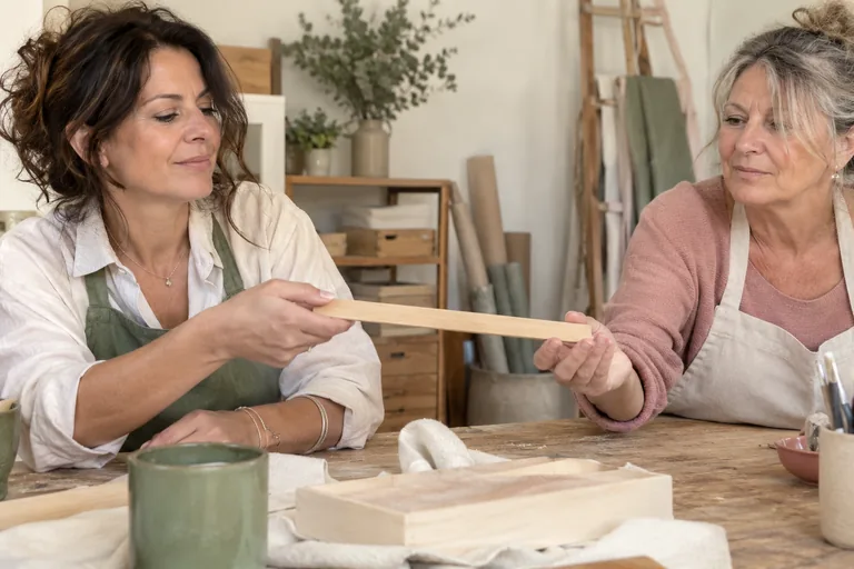 Deux femmes en tablier se passent une baguette plate en bois au-dessus d’une table.